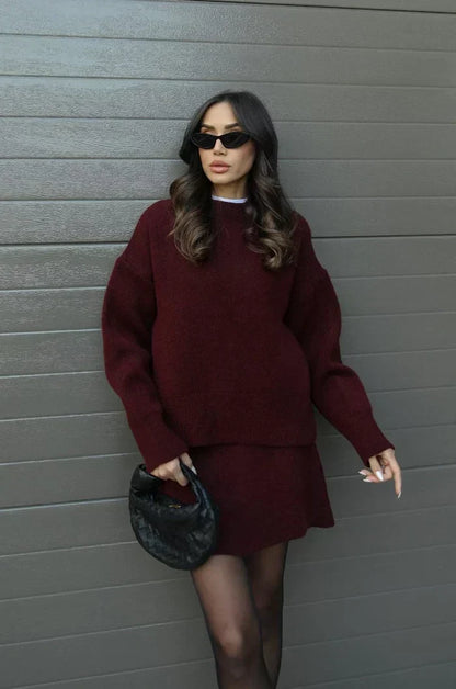 Model in weinrotem Strick-Set aus Pullover und Rock, vor einer grauen Wand stehend, schwarze Handtasche in der Hand. 1