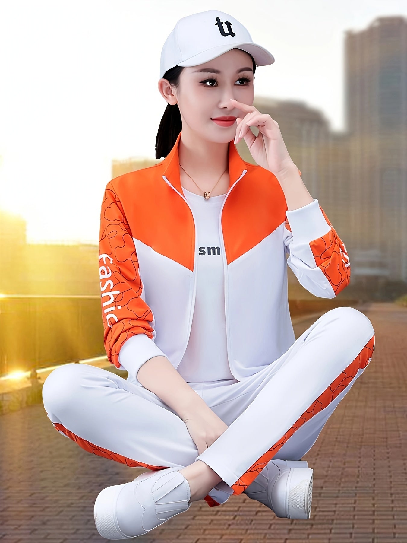 Frau in weißem und orangefarbenem Sportanzug, Baseballkappe, sitzt im Freien, Stadt im Hintergrund.