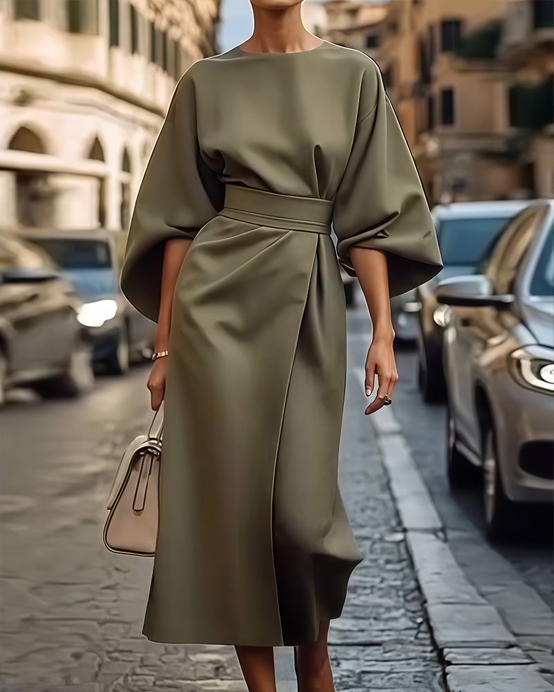 Oliv- bis khakigrünes Wickelkleid mit weiten Fledermausärmeln und Taillengürtel, getragen auf einer Kopfsteinpflasterstraße, dazu eine helle Handtasche. 2