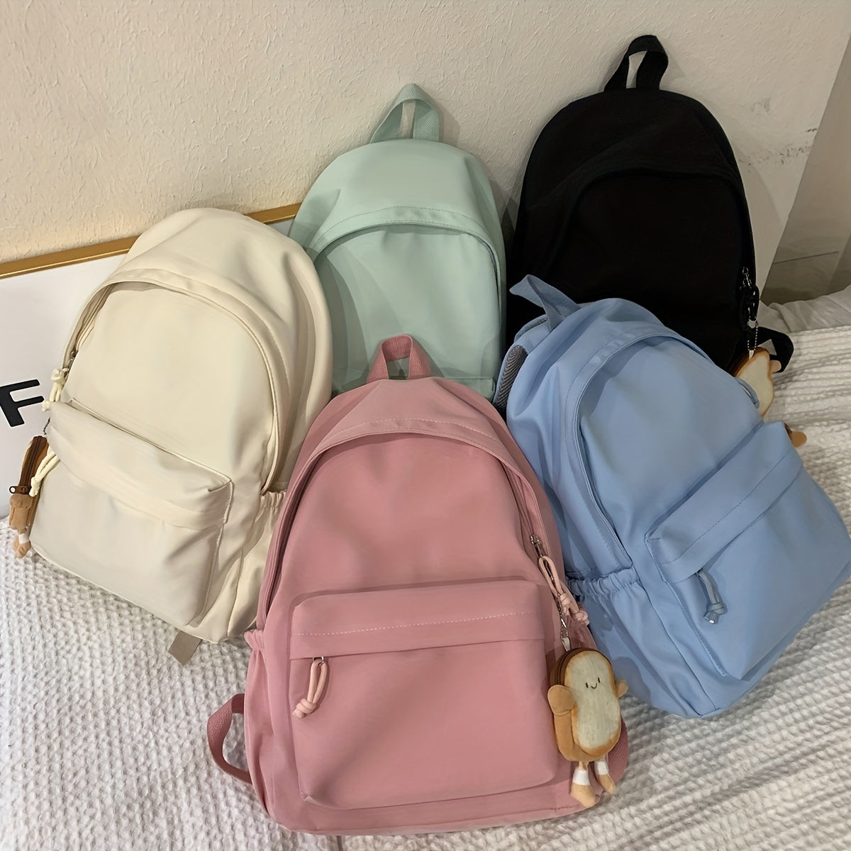 Fünf Rucksäcke in Creme, Mint, Schwarz, Rosa und Hellblau auf einem Bett, einige mit kleinem Brotscheiben-Anhänger. 8