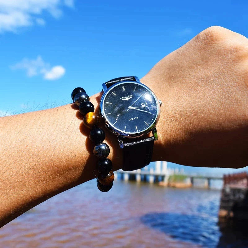 Armbanduhr mit schwarzem Zifferblatt und Lederarmband, Tigerauge-Armband, blauer Himmel.
