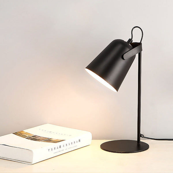 Schwarze Tischlampe mit schräg gestelltem Schirm und rundem Fuß, eingeschaltet auf einem hellen Tisch neben einem aufgeschlagenen Buch. 12