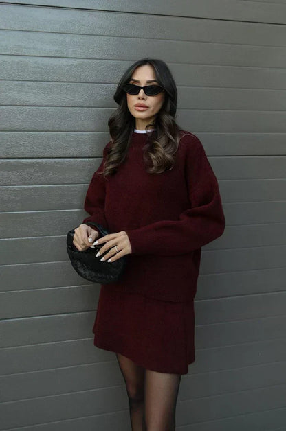 Model im weinroten Zweiteiler in leicht seitlicher Pose, hält eine kleine schwarze Tasche vor dem Körper. 3