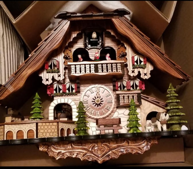 Kuckucksuhr im geöffneten Karton mit geschnitztem Unterbau, Fachwerkfassade, Tannen, Hunden und Balkon mit Figuren, Foto der Lieferung. 14
