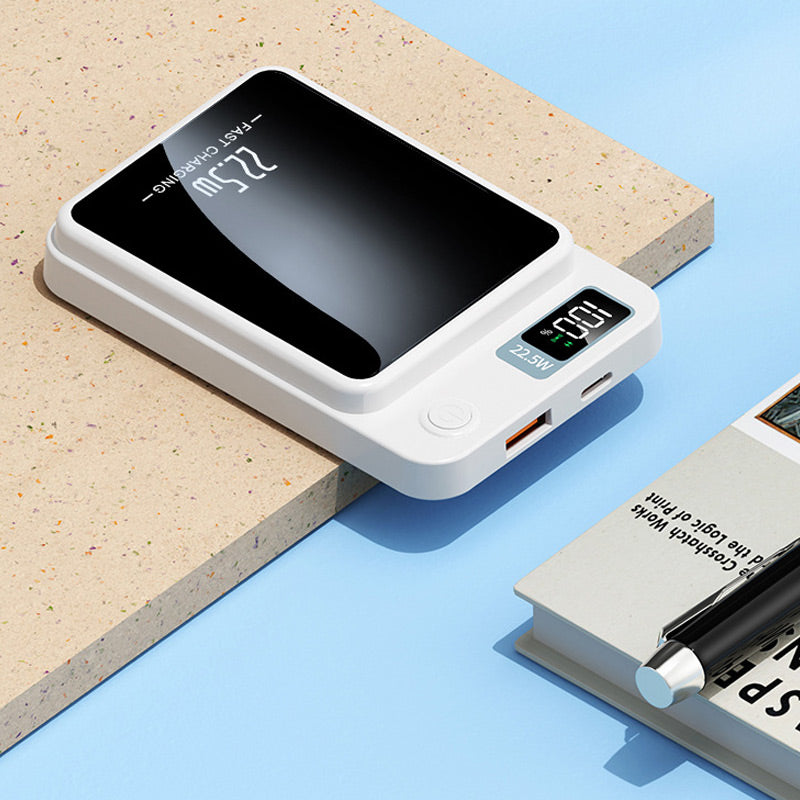 Weiße Powerbank liegt flach auf einer Kante, mit Blick auf Display, Einschalttaste, USB-A- und USB-C-Anschluss, daneben ein Buch mit Stift. 6