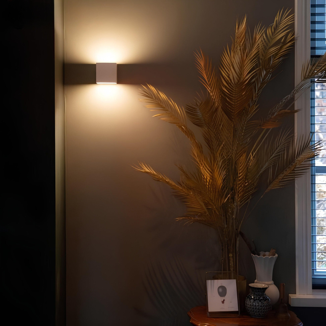 Weiße Würfelleuchte an dunkler Wand neben einer Vase mit goldenen Palmwedeln und einem Fenster mit Jalousie. 4