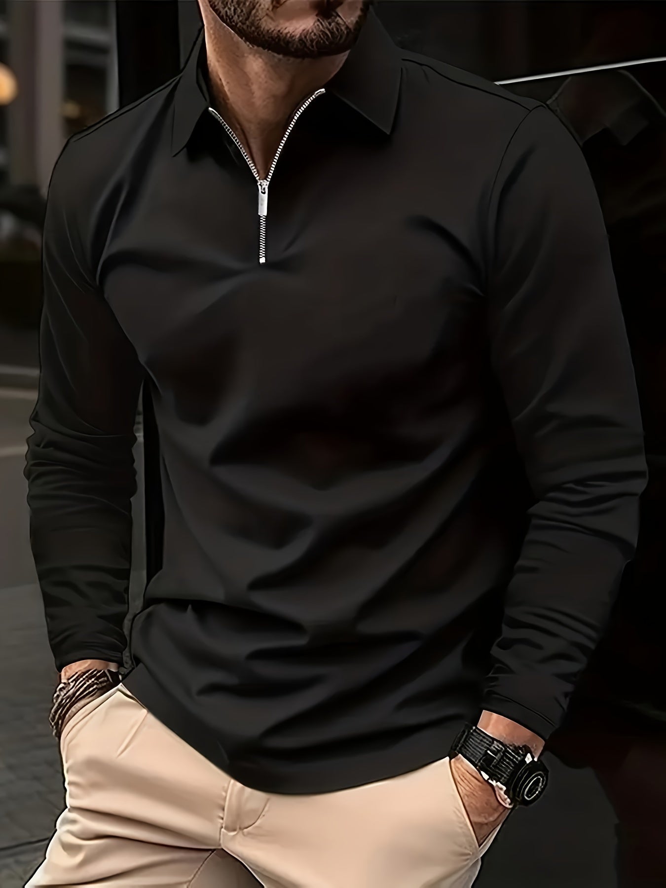 Mann in schwarzem Langarmshirt mit Reißverschluss, beige Hose, Armbanduhr, urbaner Hintergrund.