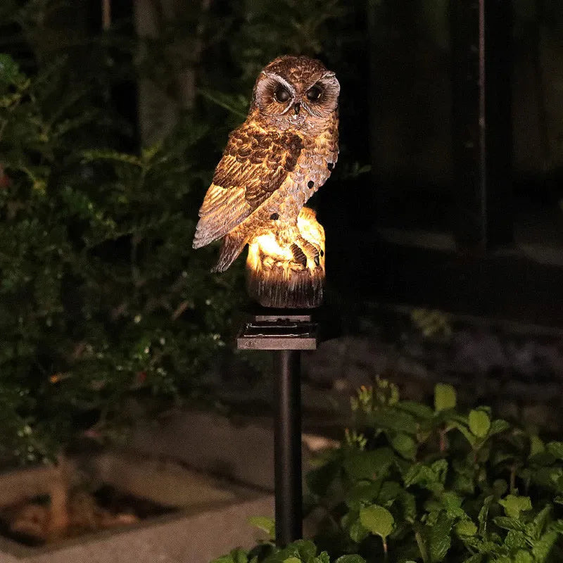 Braune Eulenleuchte leuchtet nachts warm auf einem Spieß im Beet, umgeben von dunklem Gebüsch und grünen Blättern. 4
