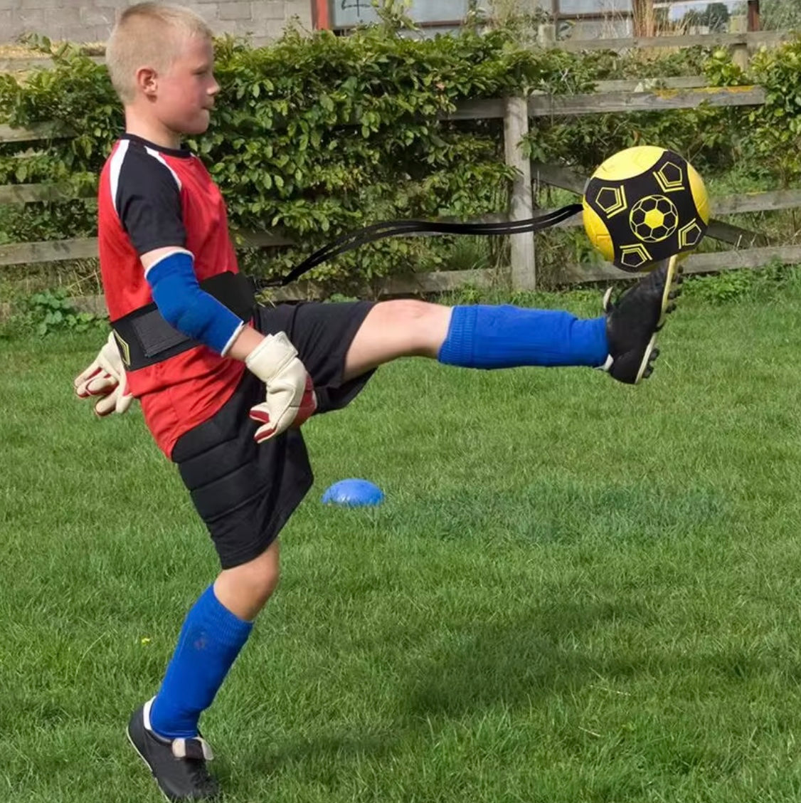 Junge trainiert mit gelbem Fußballtrainer, trägt rote Sportkleidung und blaue Stutzen.