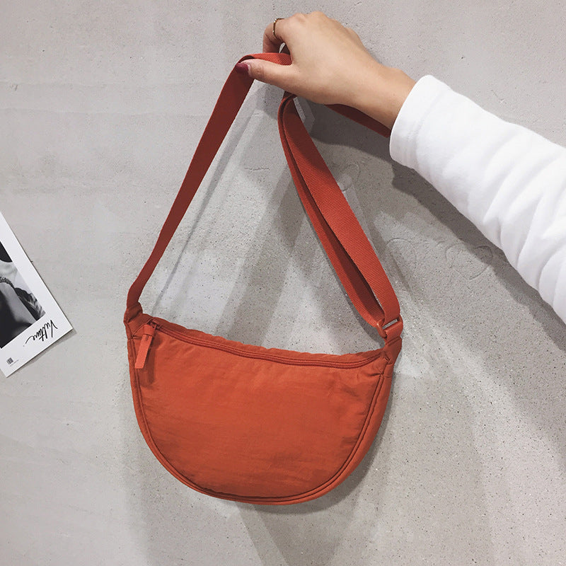 Orange Umhängetasche aus Stoff, halbmondförmig, mit Reißverschluss, an grauer Wand gehalten.