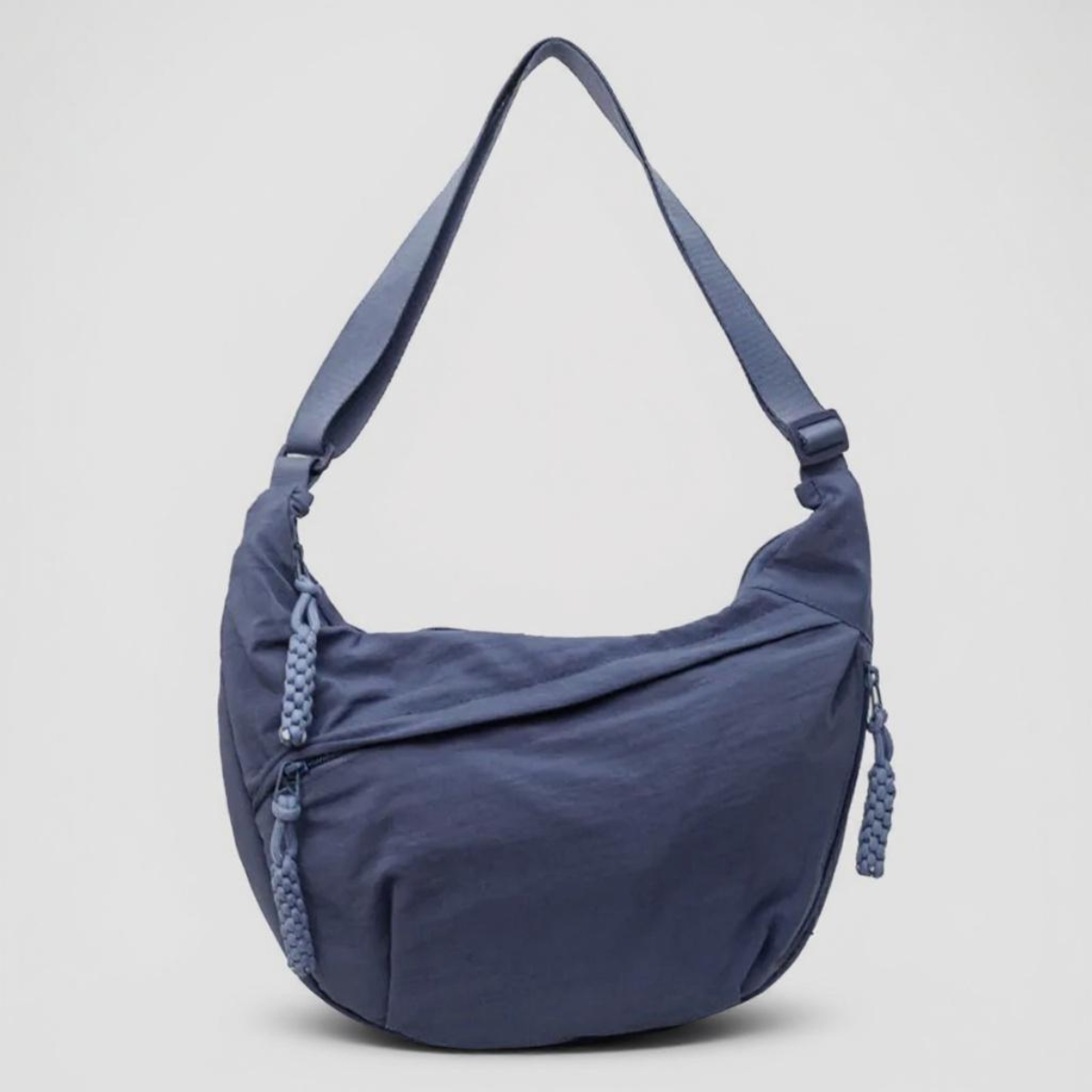 Halbmondförmige blaue Tasche mit breitem Riemen und geflochtenen Reißverschlussanhängern, frontal vor hellgrauem Hintergrund fotografiert. 1