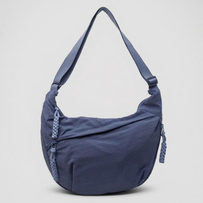 Halbmondförmige blaue Tasche mit breitem Riemen und geflochtenen Reißverschlussanhängern, frontal vor hellgrauem Hintergrund fotografiert. 1