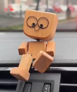 Holzfigur mit großen Drahtaugen und seitlich geneigtem Kopf sitzt auf einem Autoarmaturenbrett, ein Bein baumelt herab. 6