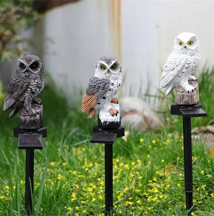 Drei Eulenfiguren in Braun, Braun-Weiß und Weiß stehen tagsüber auf Spießen in einer Blumenwiese, die Solarpanels sind gut zu erkennen. 11