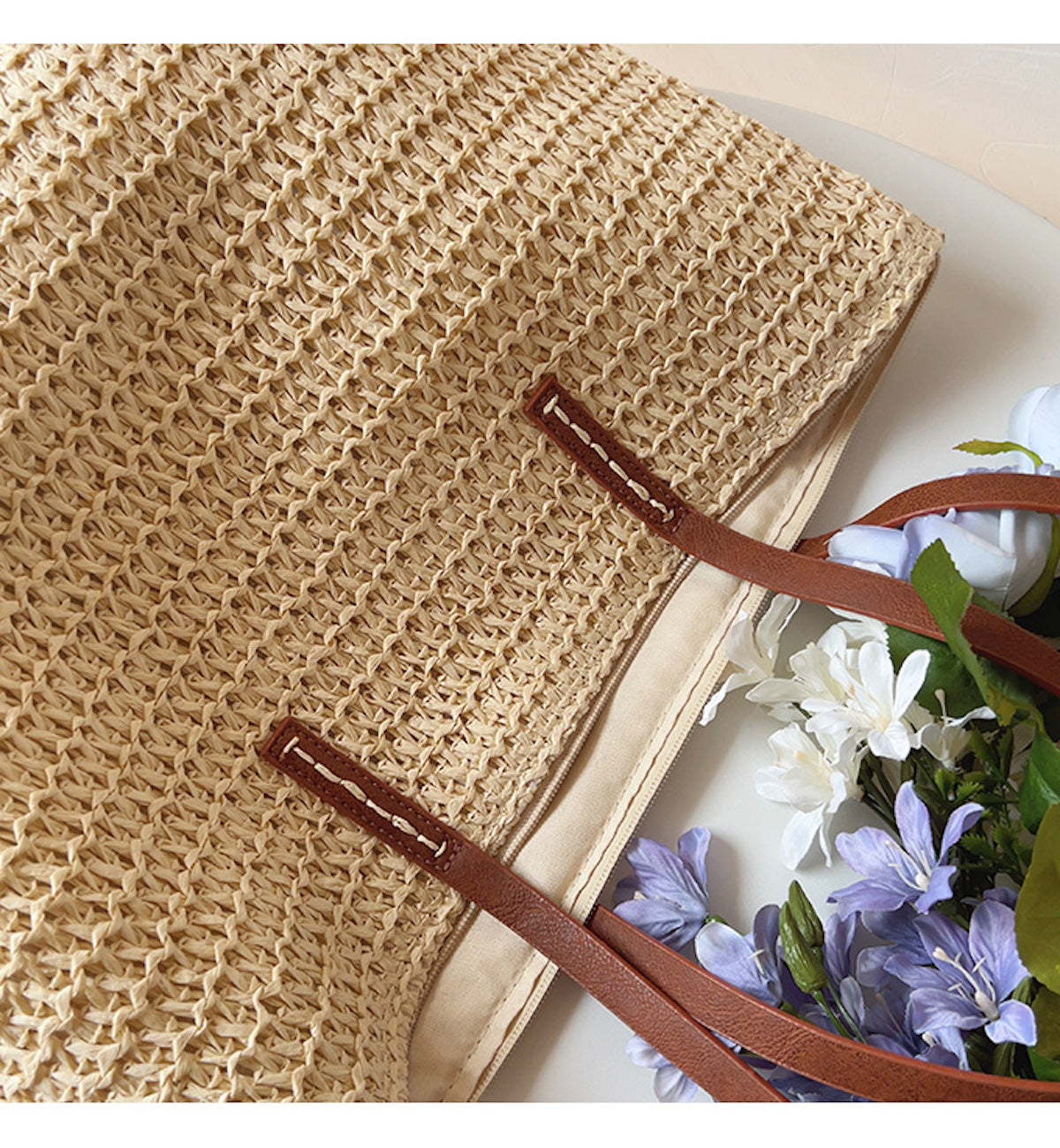 Beigefarbene Strohtasche mit braunen Ledergriffen, dekoriert mit lila und weißen Blumen.