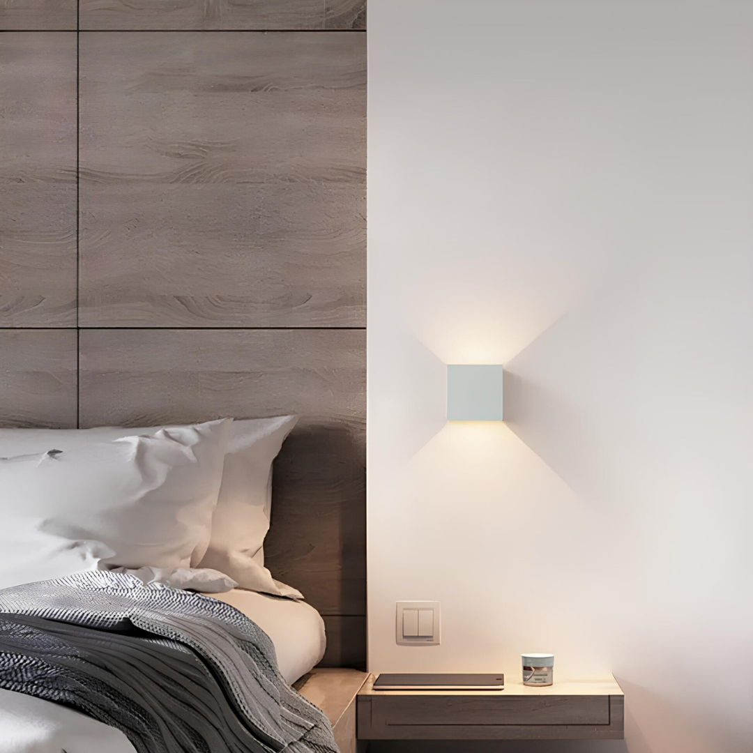 Weiße Würfelleuchte neben einem Bett mit Holzverkleidung, darunter ein schwebender Nachttisch mit Tablet und kleinem Gefäß. 6