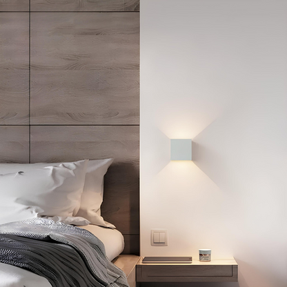 Weiße Würfelleuchte neben einem Bett mit Holzverkleidung, darunter ein schwebender Nachttisch mit Tablet und kleinem Gefäß. 6