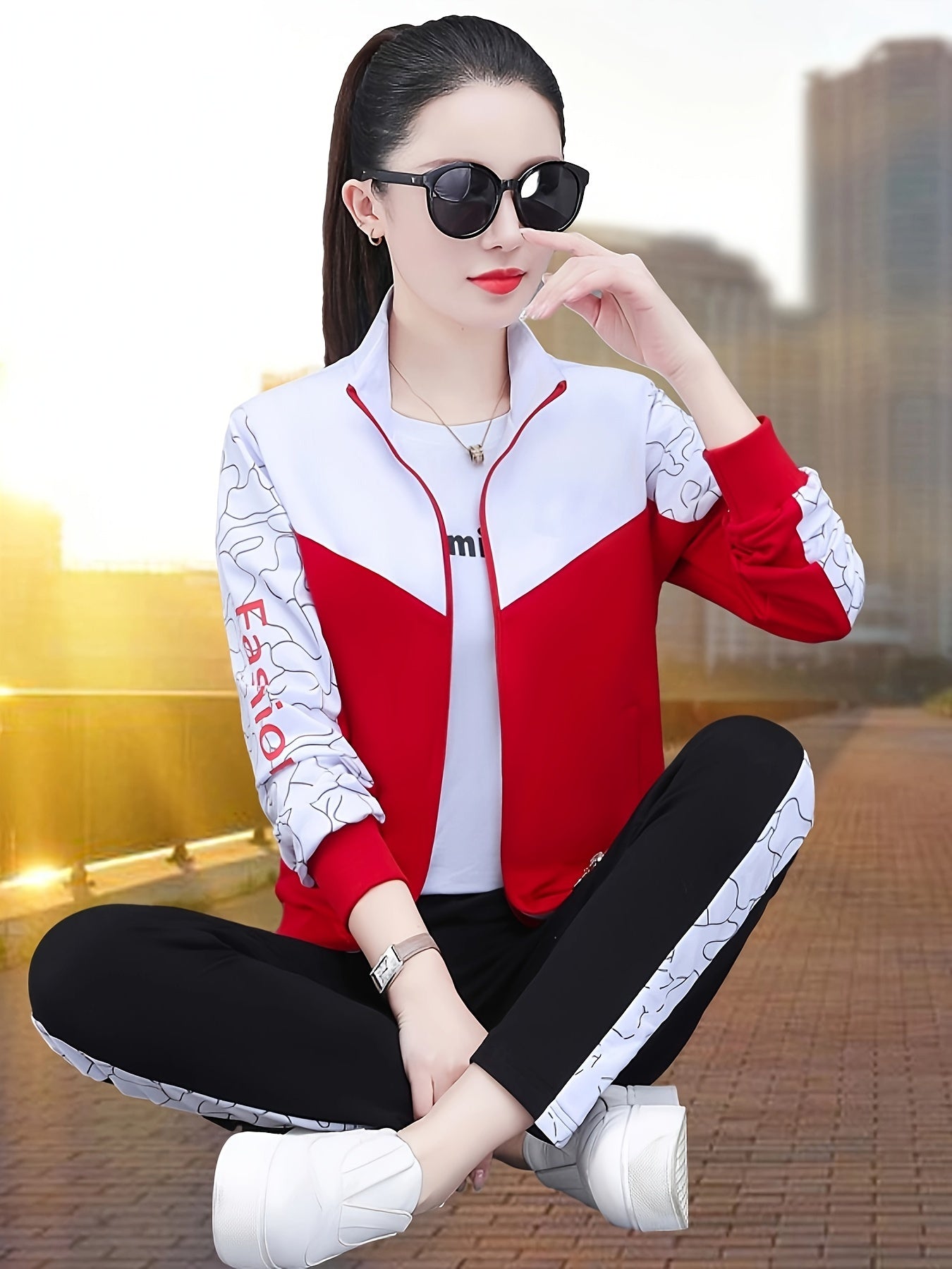 Frau in roter und weißer Sportjacke, schwarze Hose, Sonnenbrille, sitzt im Freien.