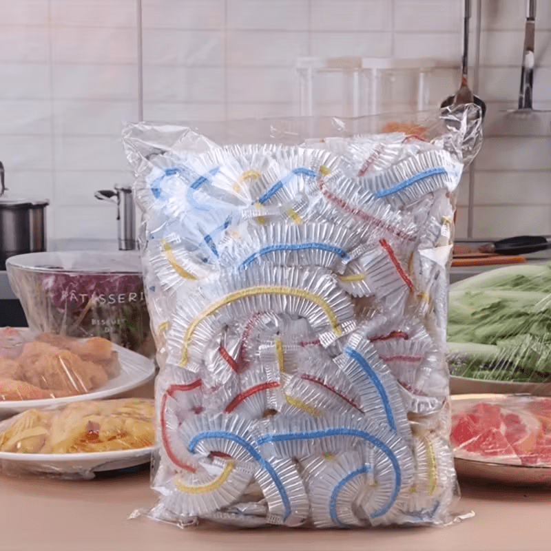 Großer Klarsichtbeutel voller Abdeckkappen mit blauen, gelben und roten Gummibändern in einer Küche, daneben abgedeckte Schalen mit Essen. 1
