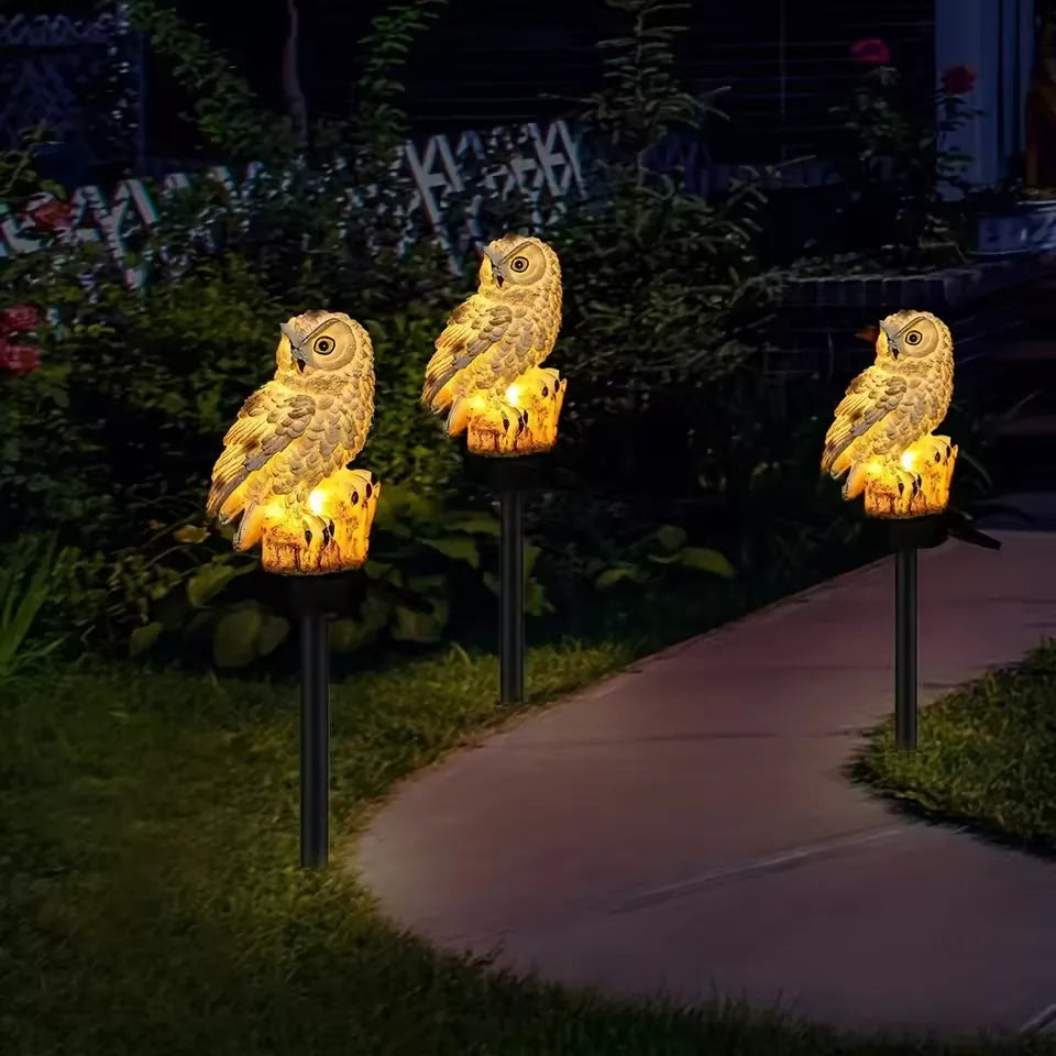 Drei warm leuchtende Eulenleuchten säumen abends einen Gartenweg, im Hintergrund Büsche mit roten Blüten. 9