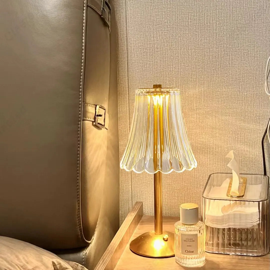 Goldene LED-Tischlampe mit Wellenschirm, kabellos, auf Nachttisch neben Parfümflasche.
