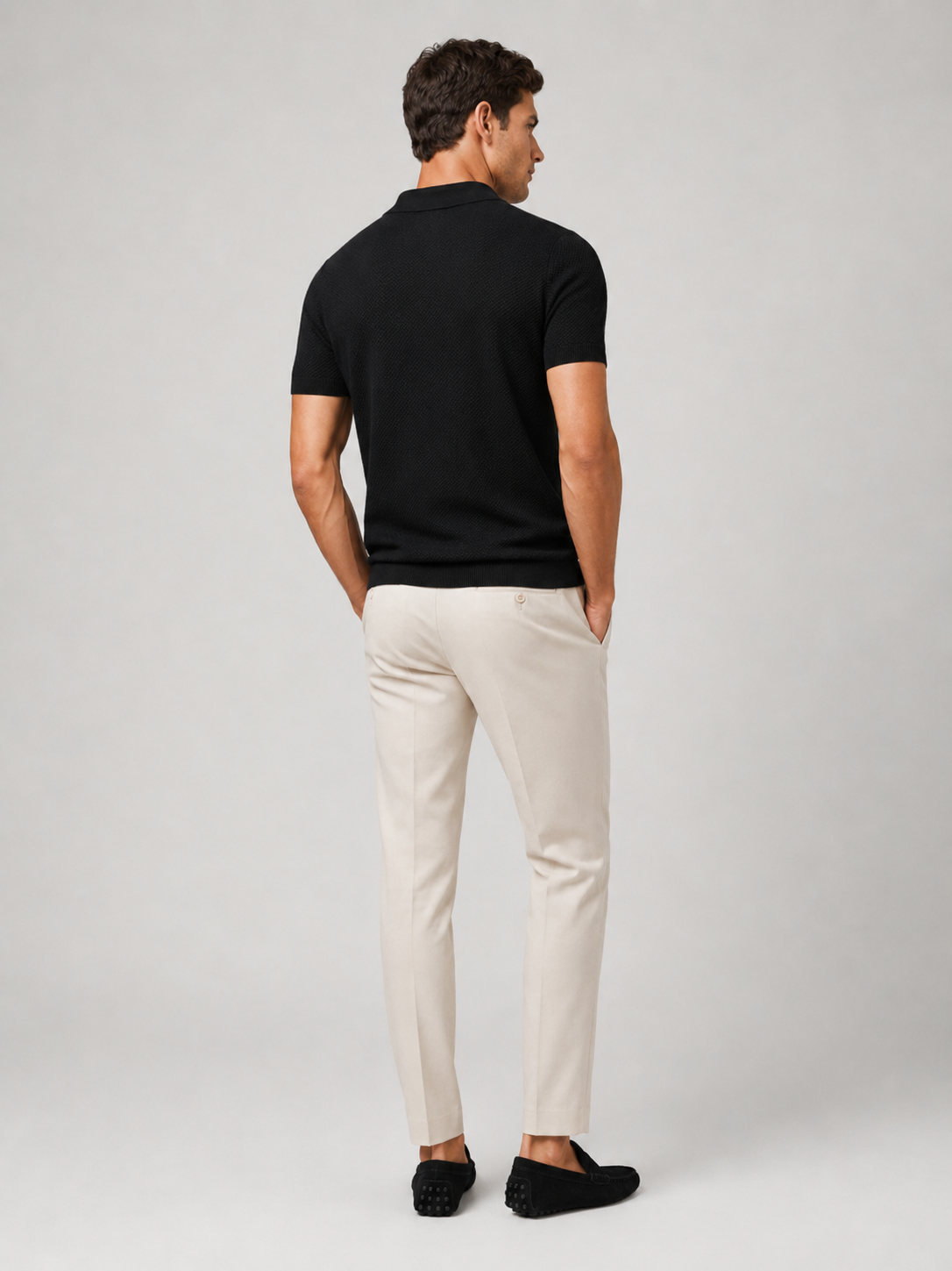 Rückansicht des schwarzen Poloshirts am Model, mit heller Hose und schwarzen Mokassins, Kopf leicht zur Seite gedreht 9