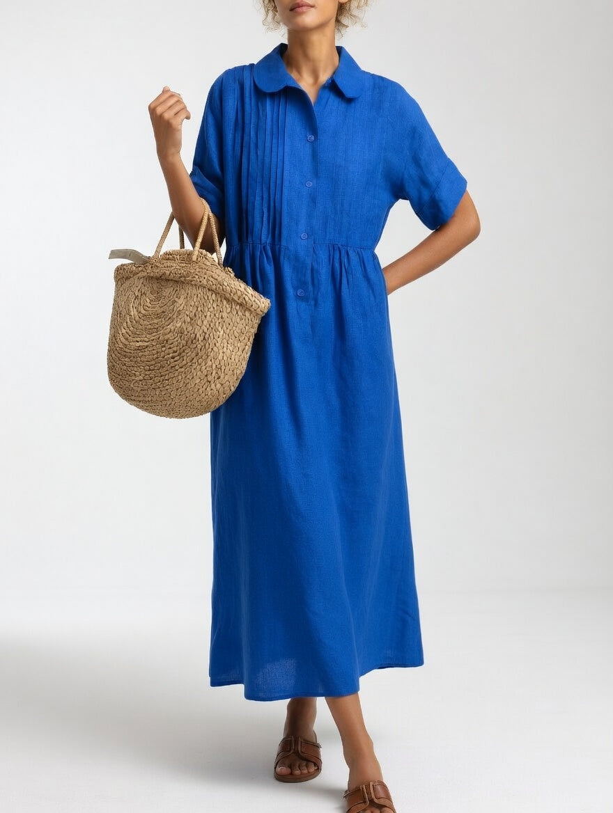Königsblaues Maxikleid in Frontansicht mit Knopfleiste und umgeschlagenen Kurzärmeln, Hand in der Hüfte, Strohtasche am Arm. 3