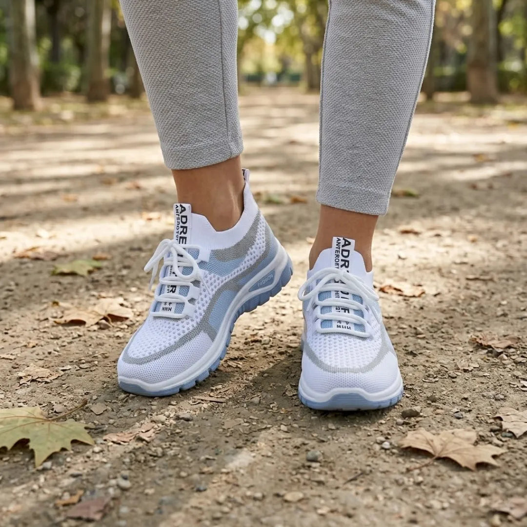 Weiße Mesh-Sneaker mit hellblauer Sohle an den Füßen einer Frau in grauer Hose auf einem Parkweg 2