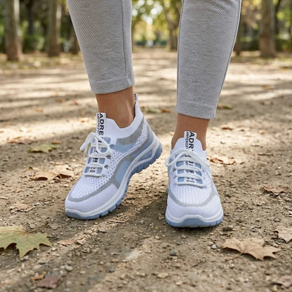 Weiße Mesh-Sneaker mit hellblauer Sohle an den Füßen einer Frau in grauer Hose auf einem Parkweg 2