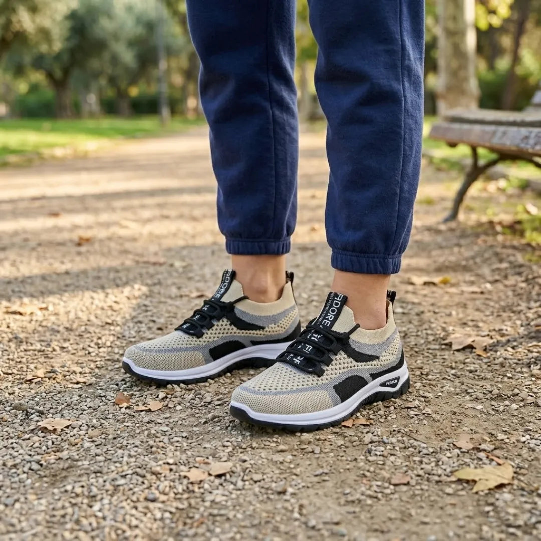 Beige Mesh-Sneaker mit schwarzen Schnürsenkeln, getragen von einer Person in dunkelblauer Jogginghose auf einem Parkweg neben einer Bank. 38