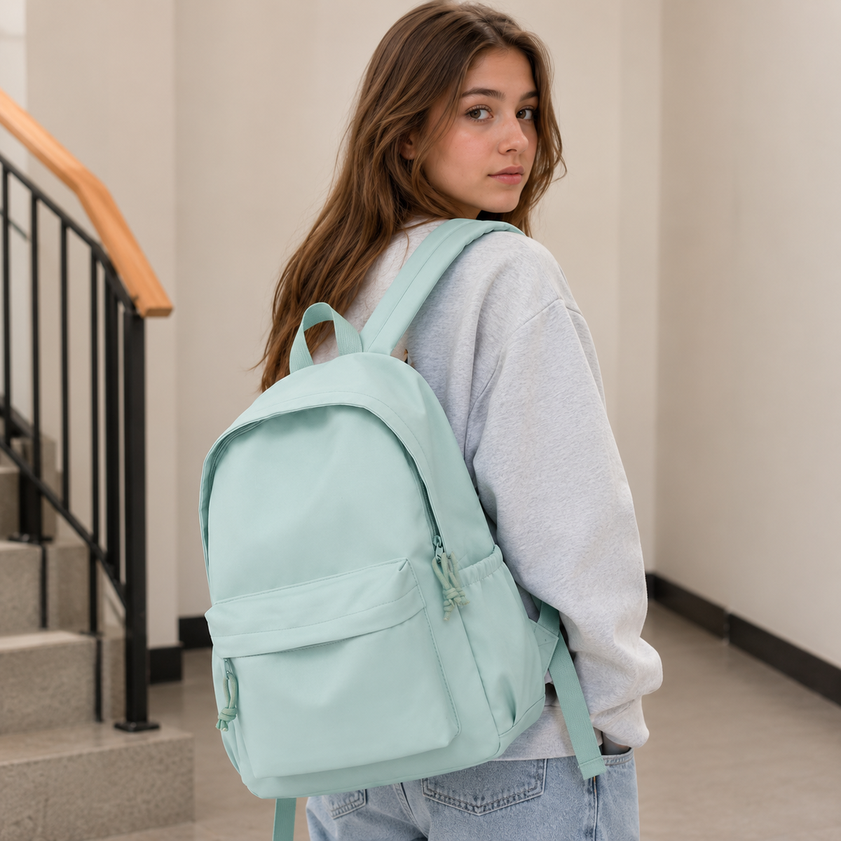 Junge Frau mit langen braunen Haaren trägt den mintgrünen Rucksack über einer Schulter, im Treppenhaus, mit grauem Pullover und Jeans. 3