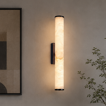Wandlampe mit marmoriertem Zylinder und schwarzen Kappen leuchtet warm an einer beigen Wand neben einem Bild und einem Zweig. 3