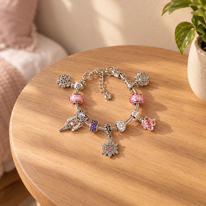 Fertig gestaltetes Charm-Armband mit rosa, violetten und klaren Strassperlen sowie Schneeflocken- und Blumenanhängern auf einem runden Holztisch neben einer Zim 1