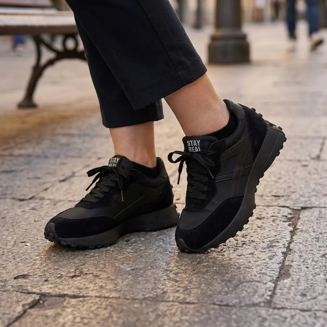 Schwarze Sneaker, getragen zu dunkler Hose auf hellem Steinpflaster, schwarze Schnürsenkel 7