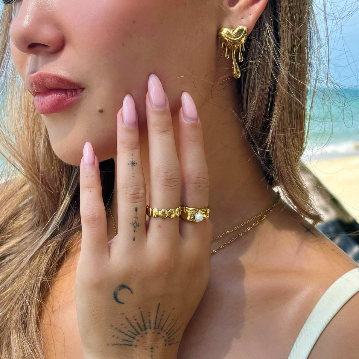 Nahaufnahme einer Frau am Strand, die einen goldfarbenen Tropfen-Herz-Ohrring trägt, dazu goldene Ringe und Ketten. 4
