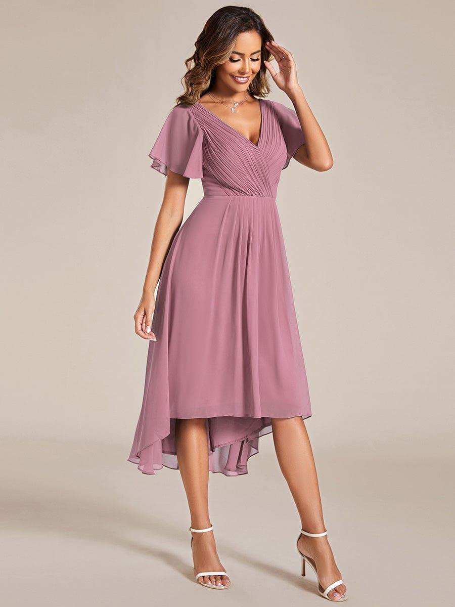 Mauvefarbenes Kleid mit Vokuhila-Saum und Flatterärmeln in Ganzkörperansicht von vorn, Model mit Hand am Kopf und weißen Sandaletten. 62