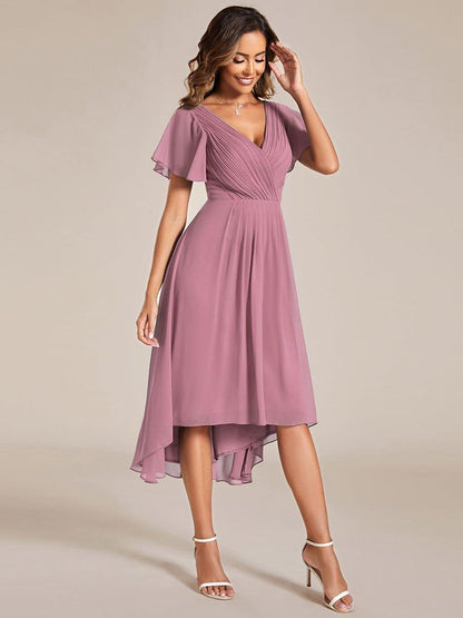 Mauvefarbenes Kleid mit Vokuhila-Saum und Flatterärmeln in Ganzkörperansicht von vorn, Model mit Hand am Kopf und weißen Sandaletten. 62