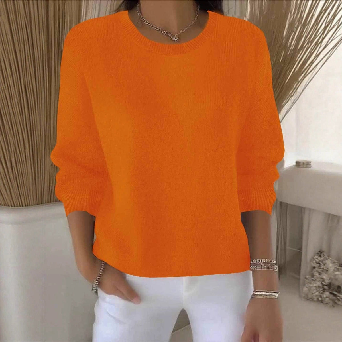 Frontansicht des orangefarbenen Strickpullovers mit Rundhalsausschnitt, getragen zu weißer Hose vor hellem Interieur 11