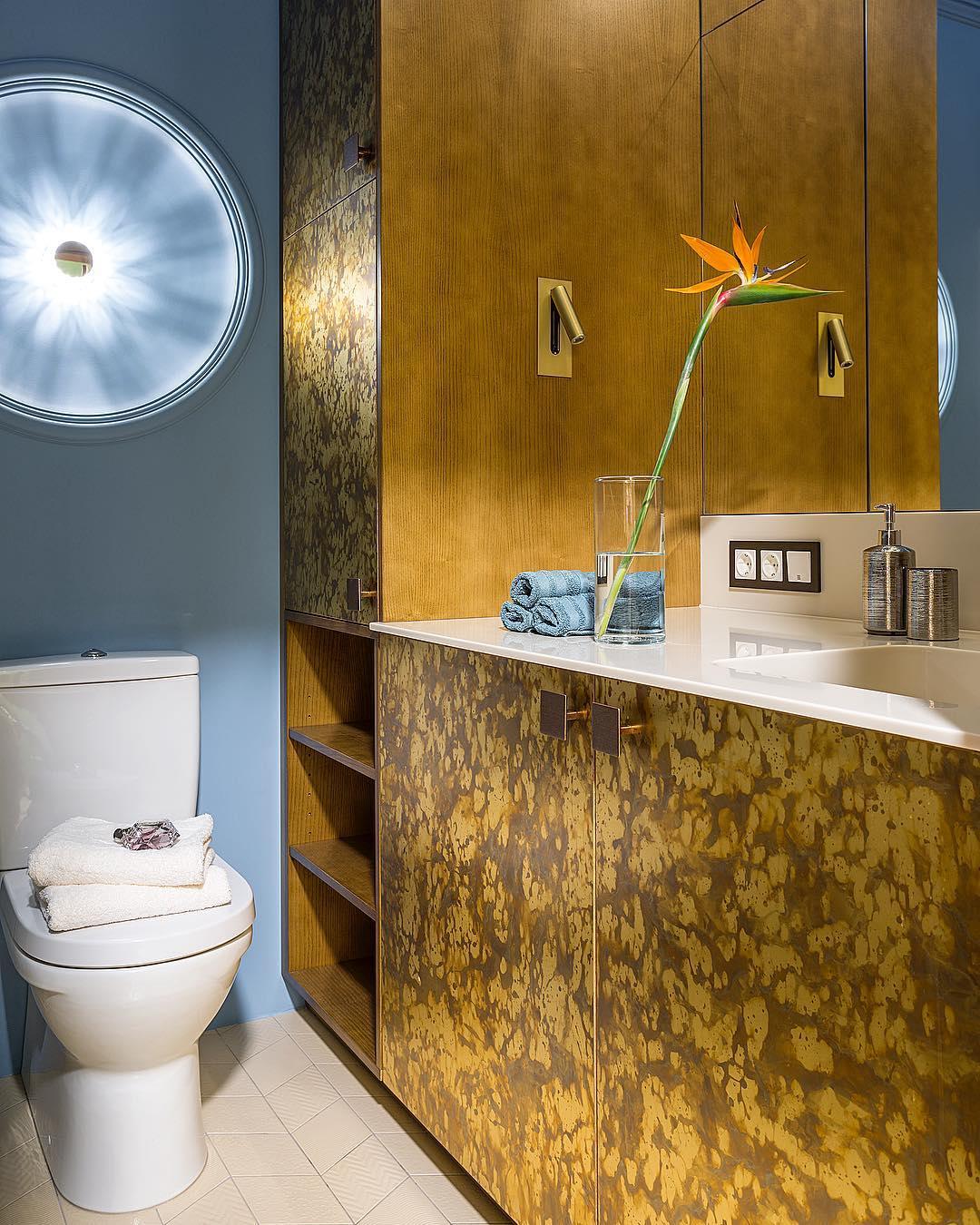 Badezimmer mit goldfarben gemusterten Schranktüren, zwei kleinen Wandleuchten am Spiegel und Strelitzie in einer Vase 10