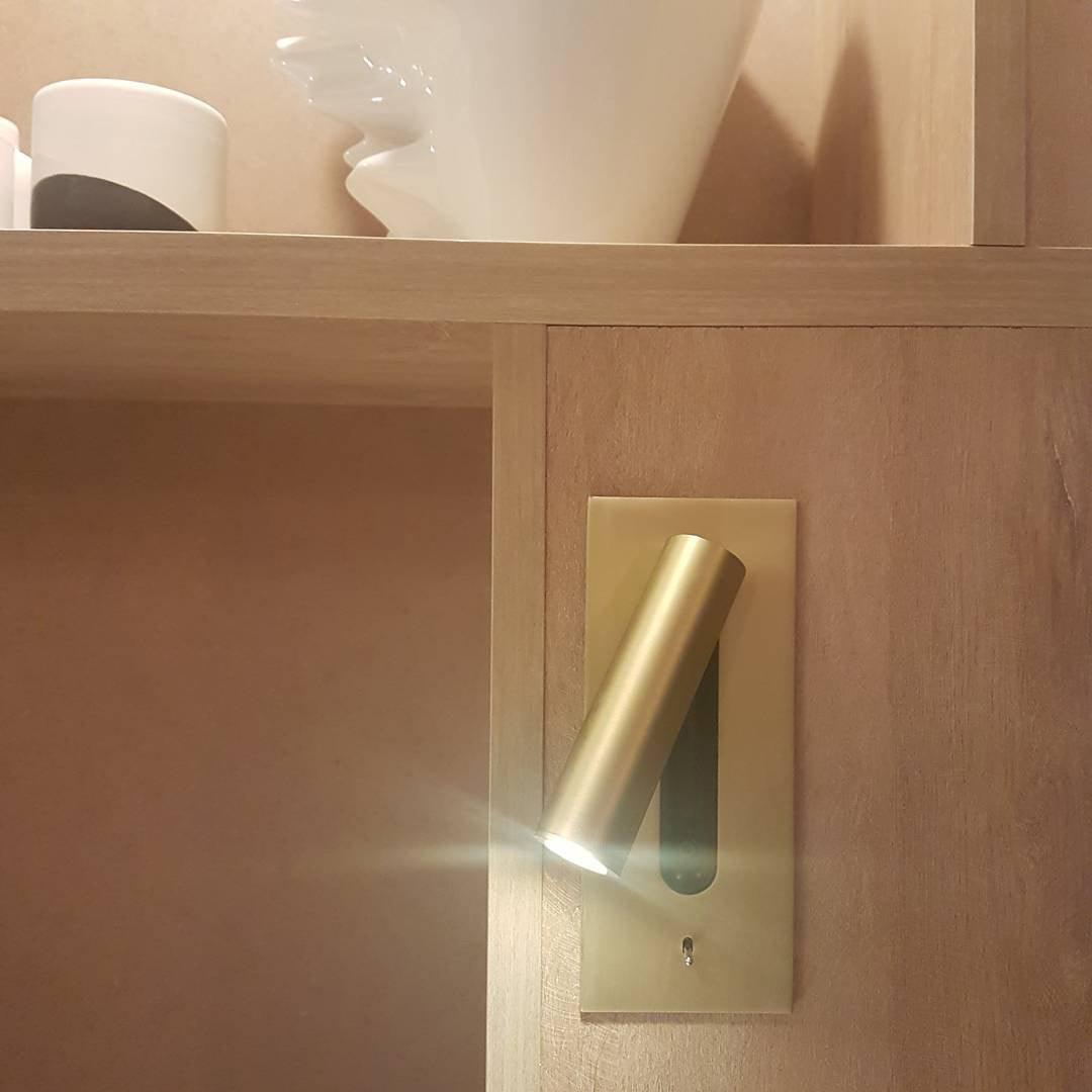 Goldfarbene Wandleuchte an einem Holzregal, der Zylinder ist nach unten geschwenkt und strahlt helles Licht ab 32