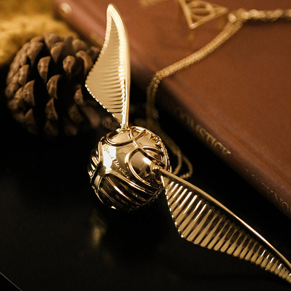 Goldener Schnatz-Anhänger mit Kette, inspiriert von Harry Potter, auf Buch und Tannenzapfen.