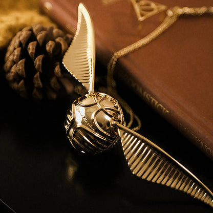 Goldener Schnatz-Anhänger mit Kette, inspiriert von Harry Potter, auf Buch und Tannenzapfen.