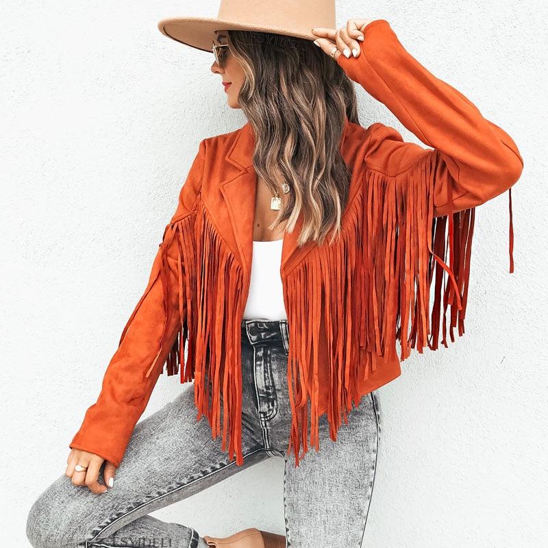 Frau in orangefarbener Fransenjacke, Jeans und Hut vor weißer Wand.