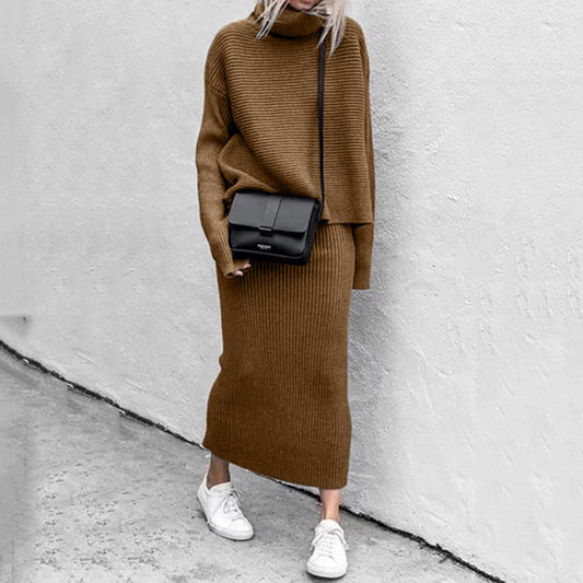 Frau in braunem Strickkleid mit schwarzer Umhängetasche und weißen Sneakers vor grauer Wand.