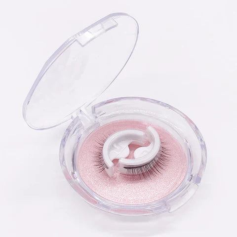 Geöffnete runde Klarsichtbox mit einem Paar Wimpern auf weißem Halter vor rosafarbenem, glitzerndem Untergrund. 1