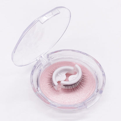 Geöffnete runde Klarsichtbox auf weißem Hintergrund, darin ein Wimpernpaar auf weißem Halter über rosa glitzerndem Einleger. 6