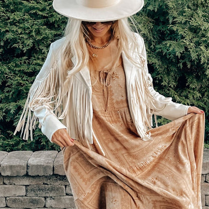 Frau in weißer Fransenjacke und beigem Kleid, Hut, Sonnenbrille, vor grüner Hecke.