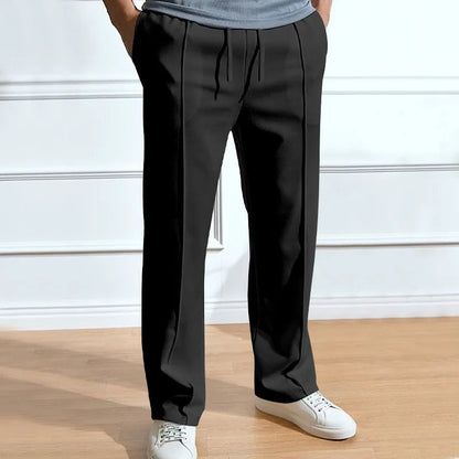 Frontansicht der Hose in Schwarz, getragen mit grauem Shirt und weißen Sneakern, Hände in den Taschen, Kordelzug und Falten sichtbar. 3