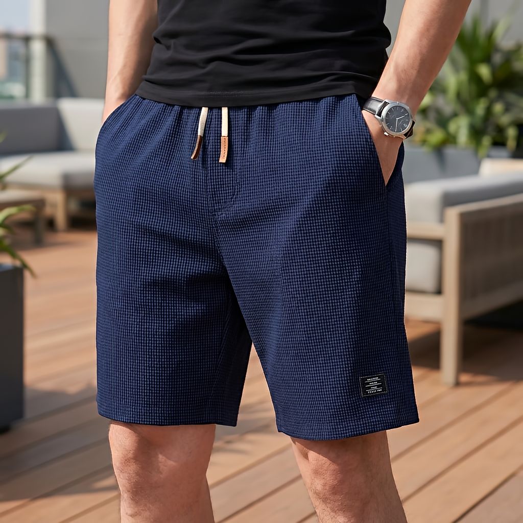 Mann in marineblauer Waffelstruktur-Short mit hellem Kordelzug und Etikett am Bein, Hände in den Taschen, auf einer Dachterrasse. 7