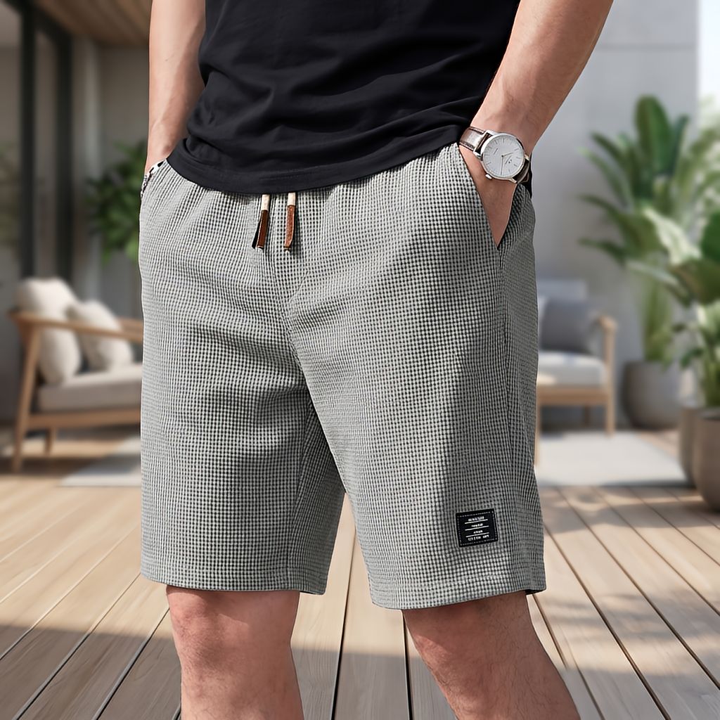 Mann in grauer Short mit feiner Waffelstruktur, Kordelzug mit Lederenden und Hand in der Tasche, auf einer Terrasse mit Pflanzen. 6